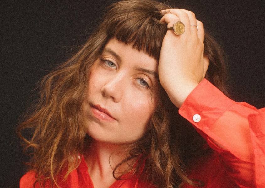 A woman wearing a red shirt with her hand rested on her head. She has a large gold coin ring on her pinky finger. She has a curly brown bob with a fringex